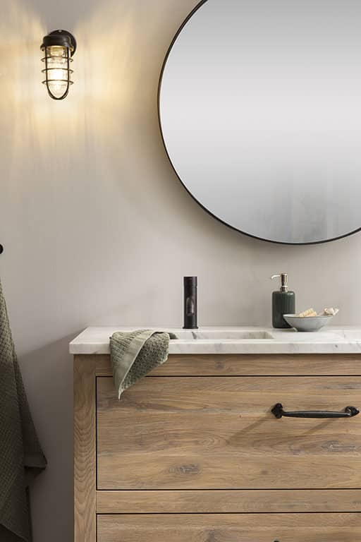 Badmöbels aus Holz mit einer wunderschönen Marmor-Waschtischplatte. Über dem Waschbecken hängt ein schwarzer Spiegel und an der Wand ist eine robuste industrielle Wandlampe befestigt. Das Holzmöbel bietet ausreichend Stauraum für Badezimmerutensilien und ist mit einem schwarzen Wasserhahn versehen. Die Marmor-Waschtischplatte und die industrielle Wandlampe verleihen dem Badezimmer eine luxuriöse und moderne Ausstrahlung.