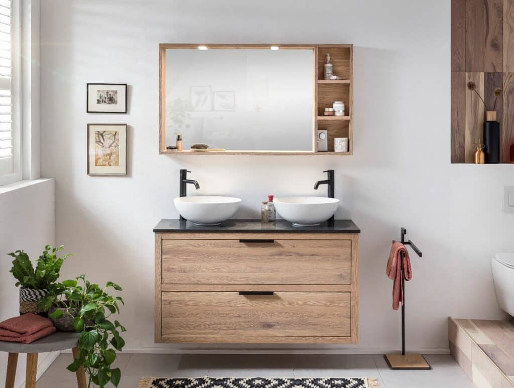 hängenden Badmöbel aus Holz mit einem Eichenspiegel und einigen grünen Pflanzen. Das Möbelstück hat ein modernes, minimalistisches Design und bietet ausreichend Stauraum für Badezimmerutensilien. Das Möbelstück ist mit einem schwarzen Wasserhahn und einer weißen Waschschüssel ausgestattet, was einen schönen Kontrast zum natürlichen Holz und den grünen Pflanzen schafft.