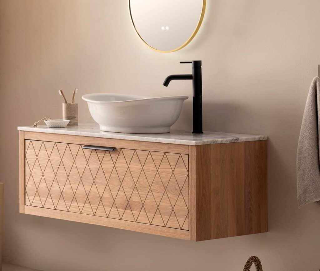 Ein schlichtes und stilvolles Badezimmer-Interieur mit einem an der Wand montierten Waschtisch aus Holz mit geometrischen Schnitzereien. Auf dem Waschtisch liegt eine Marmorplatte, auf der ein rundes Aufsatzwaschbecken und ein schwarzer Wasserhahn montiert sind. Ein beleuchteter runder Spiegel an der Wand und ein neben dem Waschtisch hängendes Handtuch vervollständigen das moderne und natürliche Design