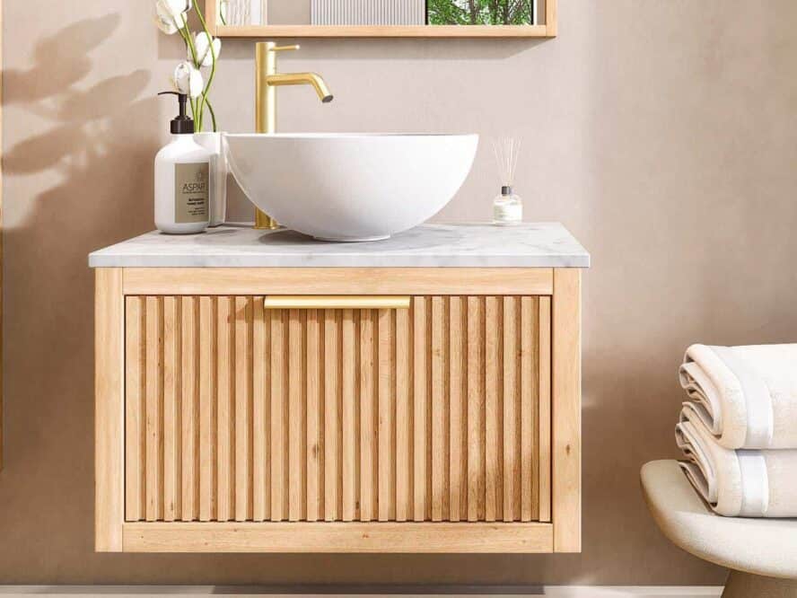 Massives Badmöbel aus Holz mit Rillenfront, 70 cm breit, hängend in einem modernen Badezimmer. Ausgestattet mit einer weißen Marmorplatte, einem Keramik-Aufsatzwaschbecken und einer goldenen, erhöhten Waschtischarmatur.“